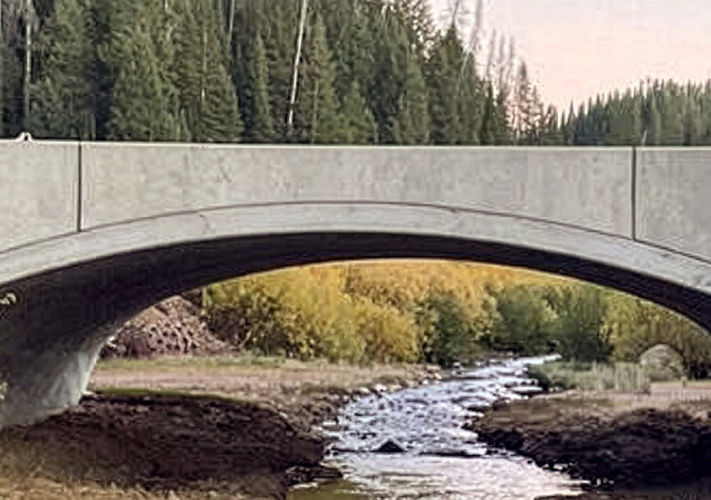 A concrete arch bridge spans a narrow stream with trees and greenery in the background.