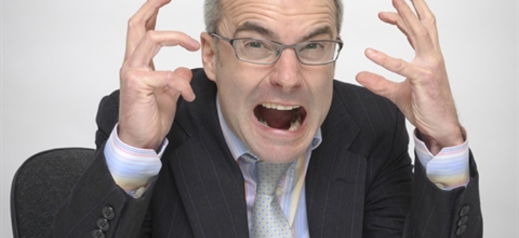 A man in a suit and tie sits at a desk, expressing frustration with his hands raised and mouth open.