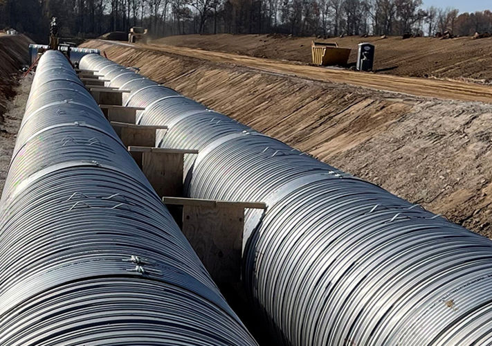 Two large metal pipes laid side by side in a trench at a construction site with machinery in the background.