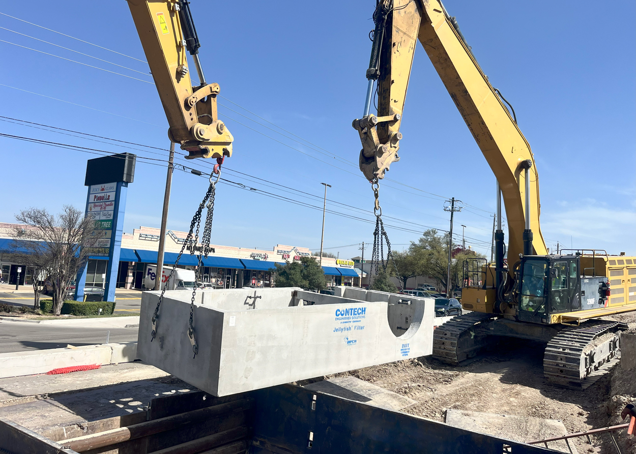 Two excavators lift a large concrete structure at a construction site beside a busy street and shops.