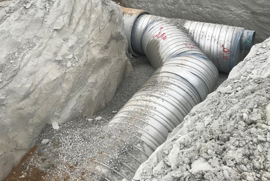 Large corrugated metal pipes installed in an excavated trench, partially covered with gravel and surrounded by soil.