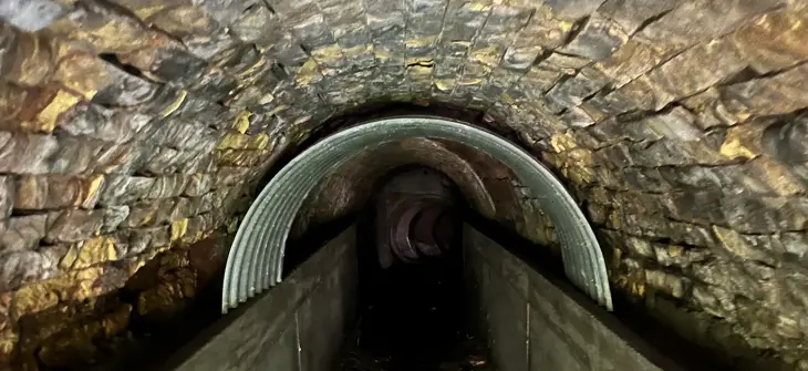 View inside a stone and concrete tunnel with an arched ceiling and corrugated metal lining partway through.