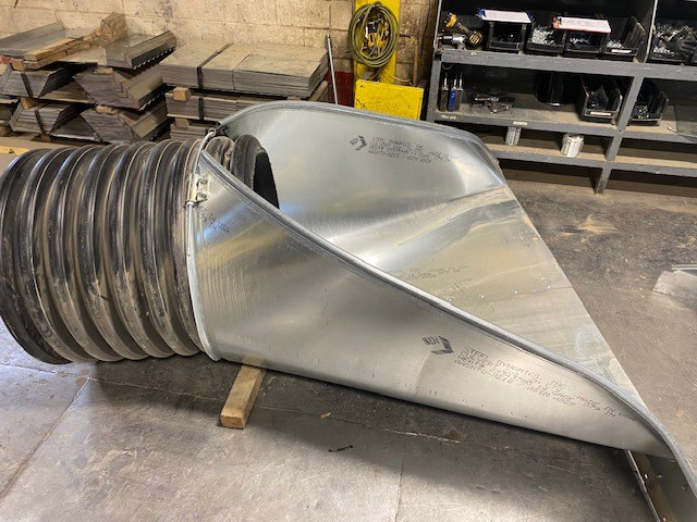 A large metal funnel with a black corrugated pipe attached, lying on a workshop floor near shelves and tools.