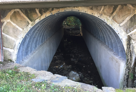 A concrete and metal culvert with a small water channel running through it, surrounded by grass and stones.