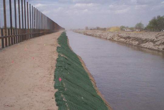 A metal fence runs alongside a canal with a green mesh-covered embankment under a partly cloudy sky.