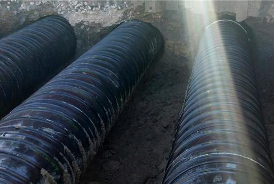 Two large black pipes buried in a trench with sunlight streaming across them.