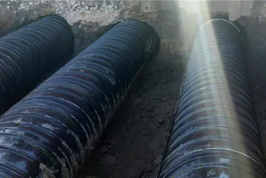 Two large black pipes buried in a trench with sunlight streaming across them.