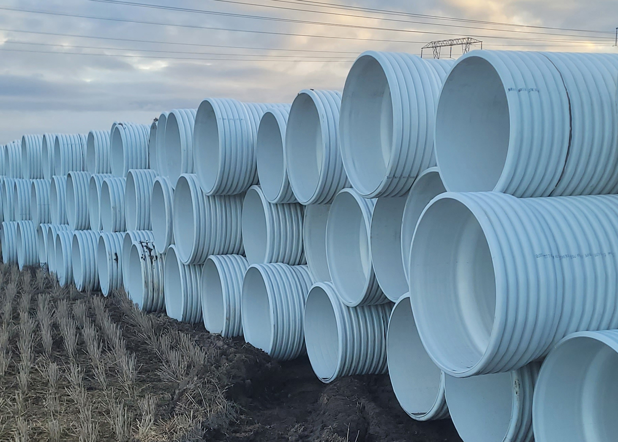 Large stacks of white, corrugated pipes are arranged in rows on a field under a cloudy sky with power lines in the background.