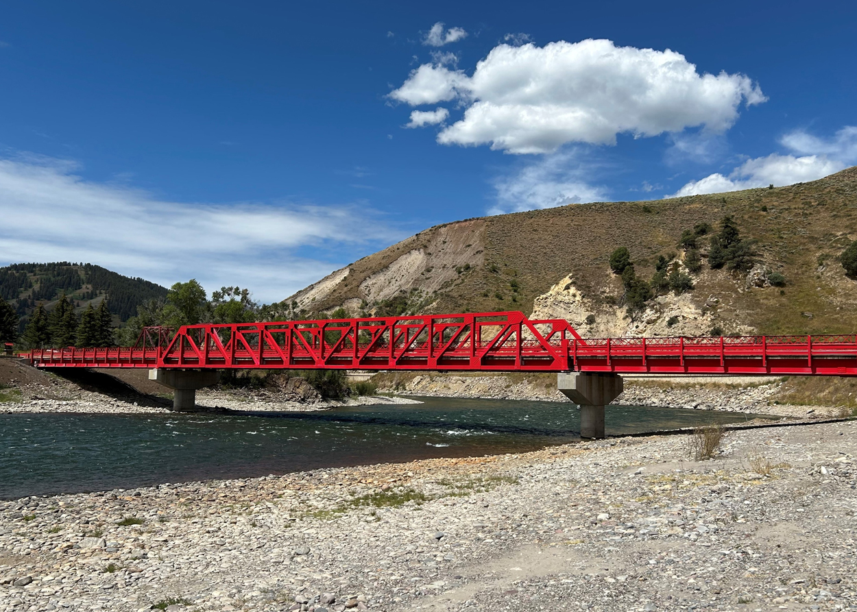 A bright red steel truss bridge spans a river with rocky banks and hills in the background under a blue sky.
