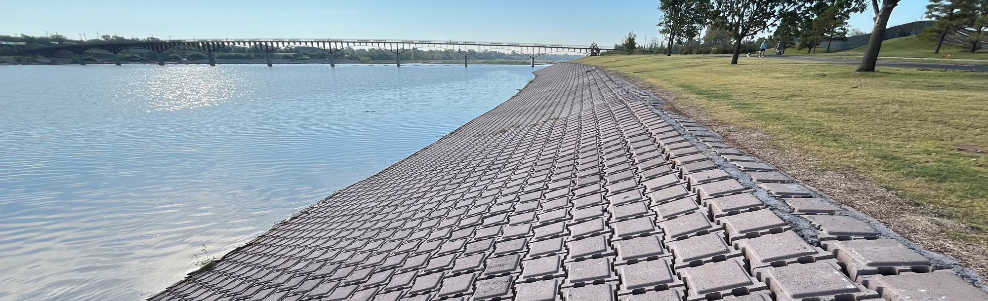 Interlocking concrete blocks line a gently sloped riverbank next to a grassy area with trees and a bridge in the background.