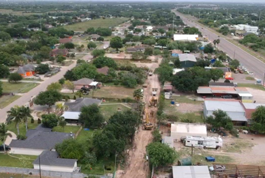 Aerial view of a residential area with road construction in progress along a street, surrounded by houses and greenery.