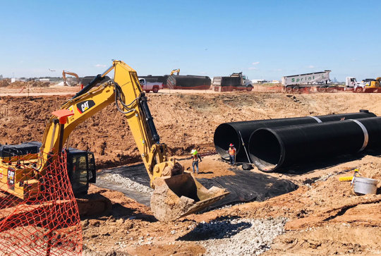 Construction site with an excavator and workers placing large black pipes into a trench under a clear blue sky.