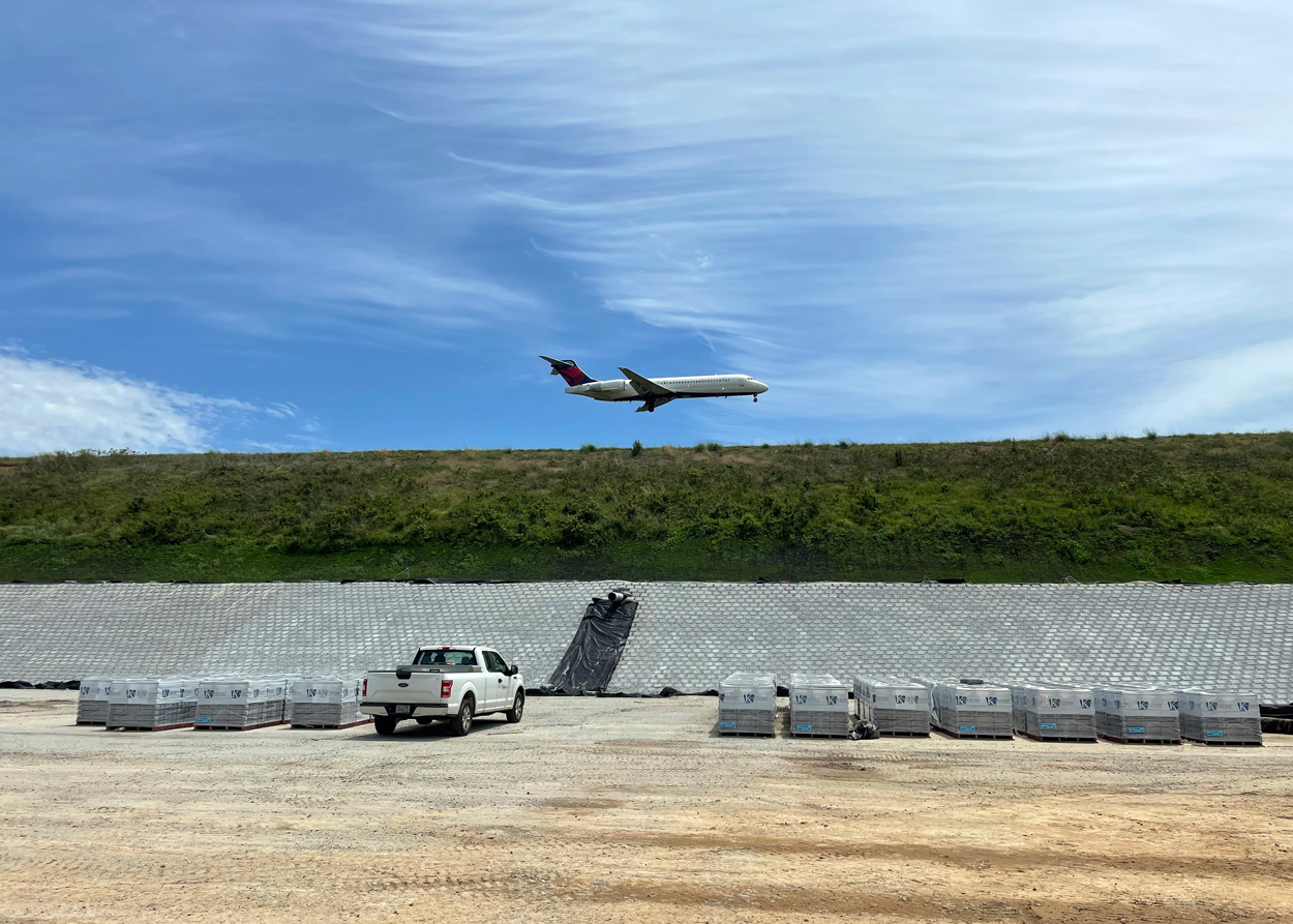 A commercial airplane flies low over a grass embankment near a construction site with a white truck and stacked materials.