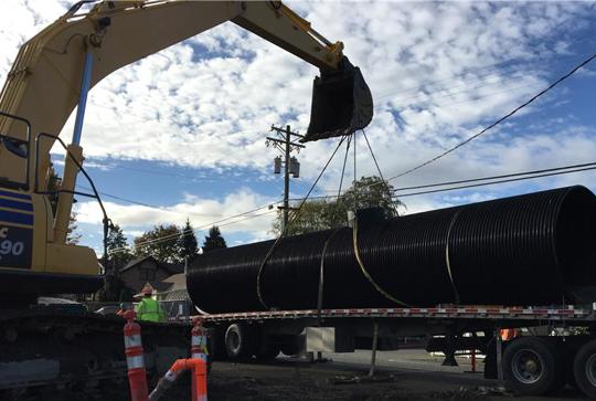 Excavator lifting a large black pipe onto a flatbed truck on a construction site, with workers and traffic cones nearby.