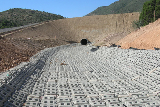 Concrete blocks line a drainage channel leading to a large pipe, surrounded by hills and sparse vegetation.
