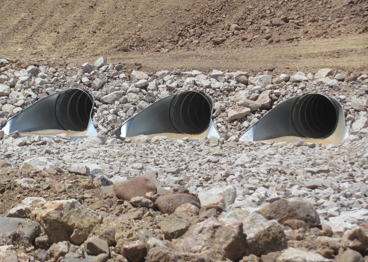 Three large black drainage pipes embedded in rocky ground at a construction or excavation site.