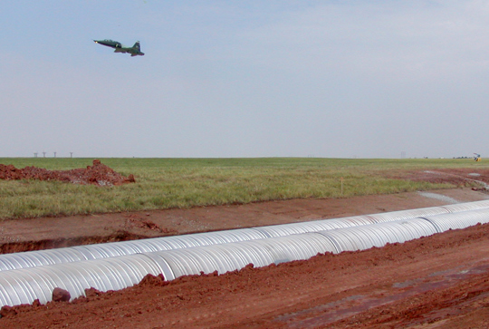 A low-flying jet soars above a green field with a construction site, featuring metallic pipes laid in the ground below.