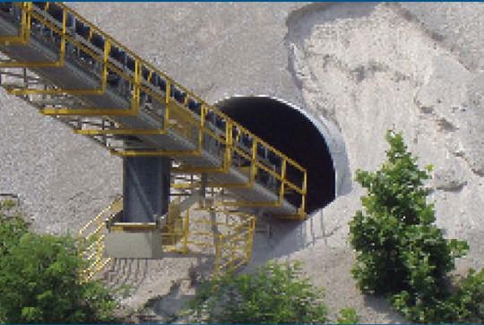 A conveyor belt transports materials into a tunnel entrance surrounded by sand and greenery.
