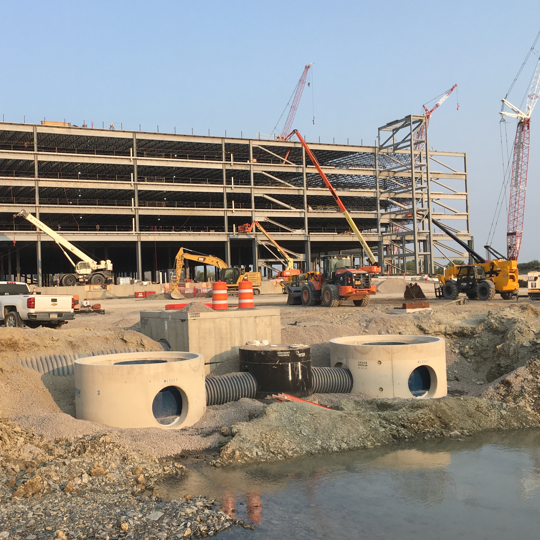 Large building under construction with cranes, vehicles, and drainage pipes visible in the foreground.