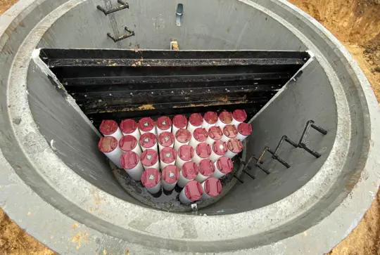 A concrete vault contains multiple red-capped cylindrical objects arranged inside, with metal steps on the inner wall.