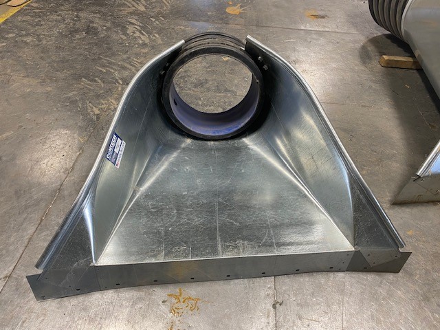A metal HVAC duct transition fitting with a round opening and flared rectangular base on a concrete floor.